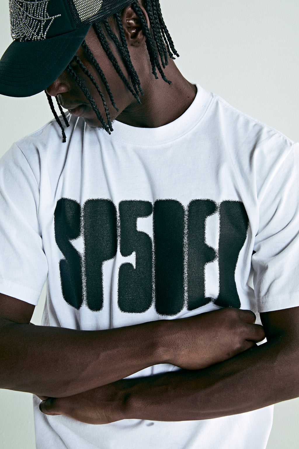 White cotton tee with bold black 'SP5DER' text print, worn by a person in a black trucker hat.