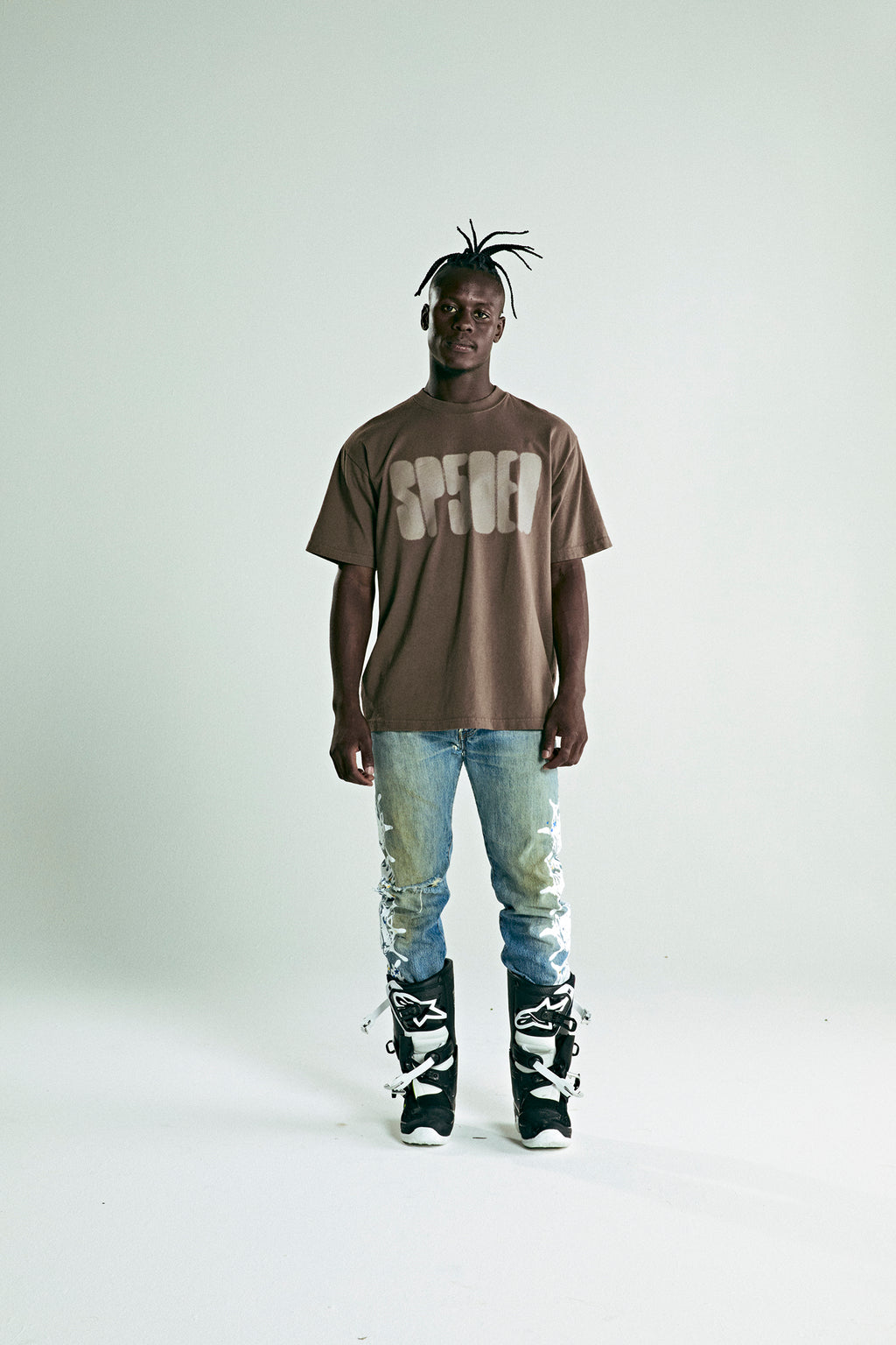 Man in a brown FOCUSED LOGO TEE with "SP5DER" text, distressed jeans, and black and white boots.