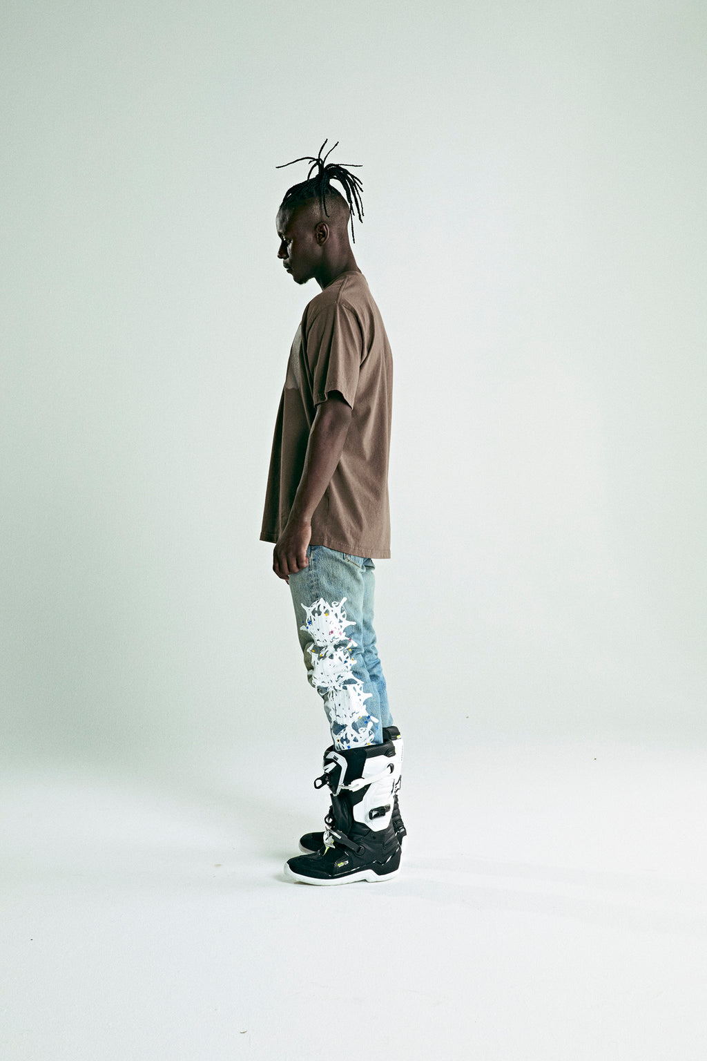 Man in brown cotton tee with 'SP5DER' text, light wash jeans with white floral print, and black boots.