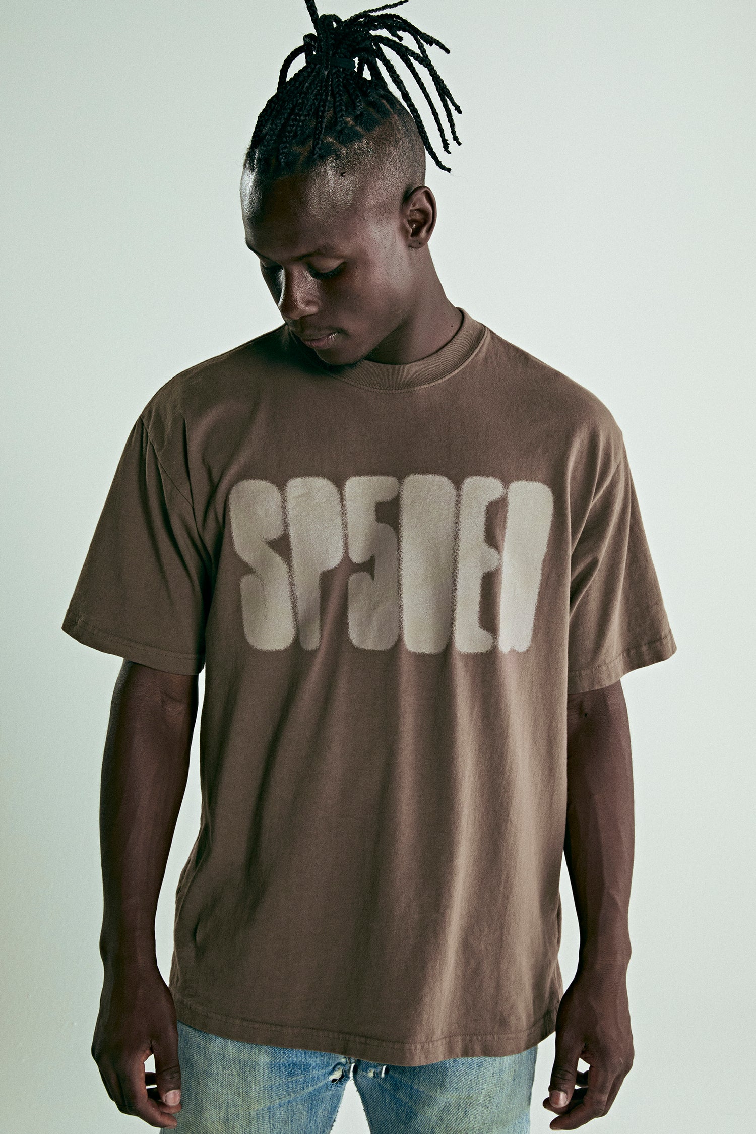 Brown cotton tee with "SP5DER" text print on front, worn by a man.