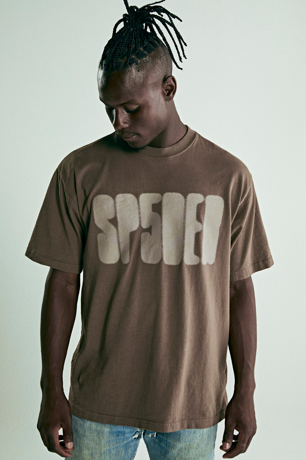 Brown cotton tee with "SP5DER" text print on front, worn by a man.