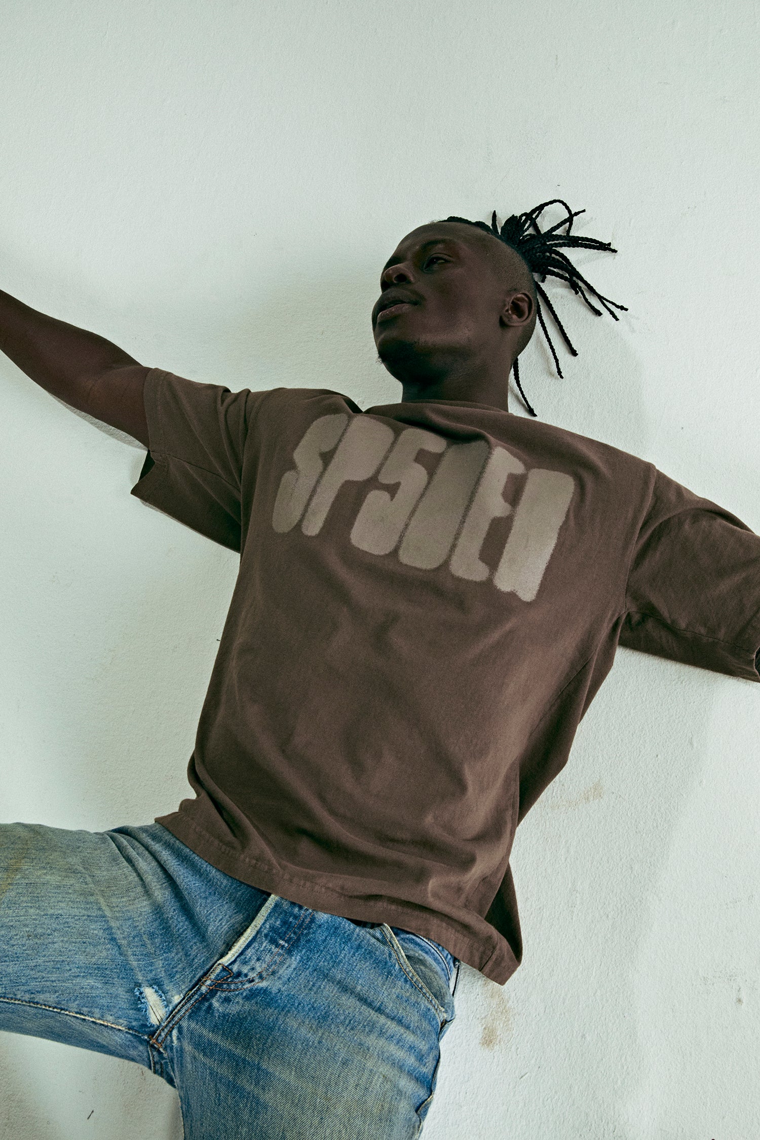 Man in a brown cotton SP5DER logo tee and distressed blue jeans.