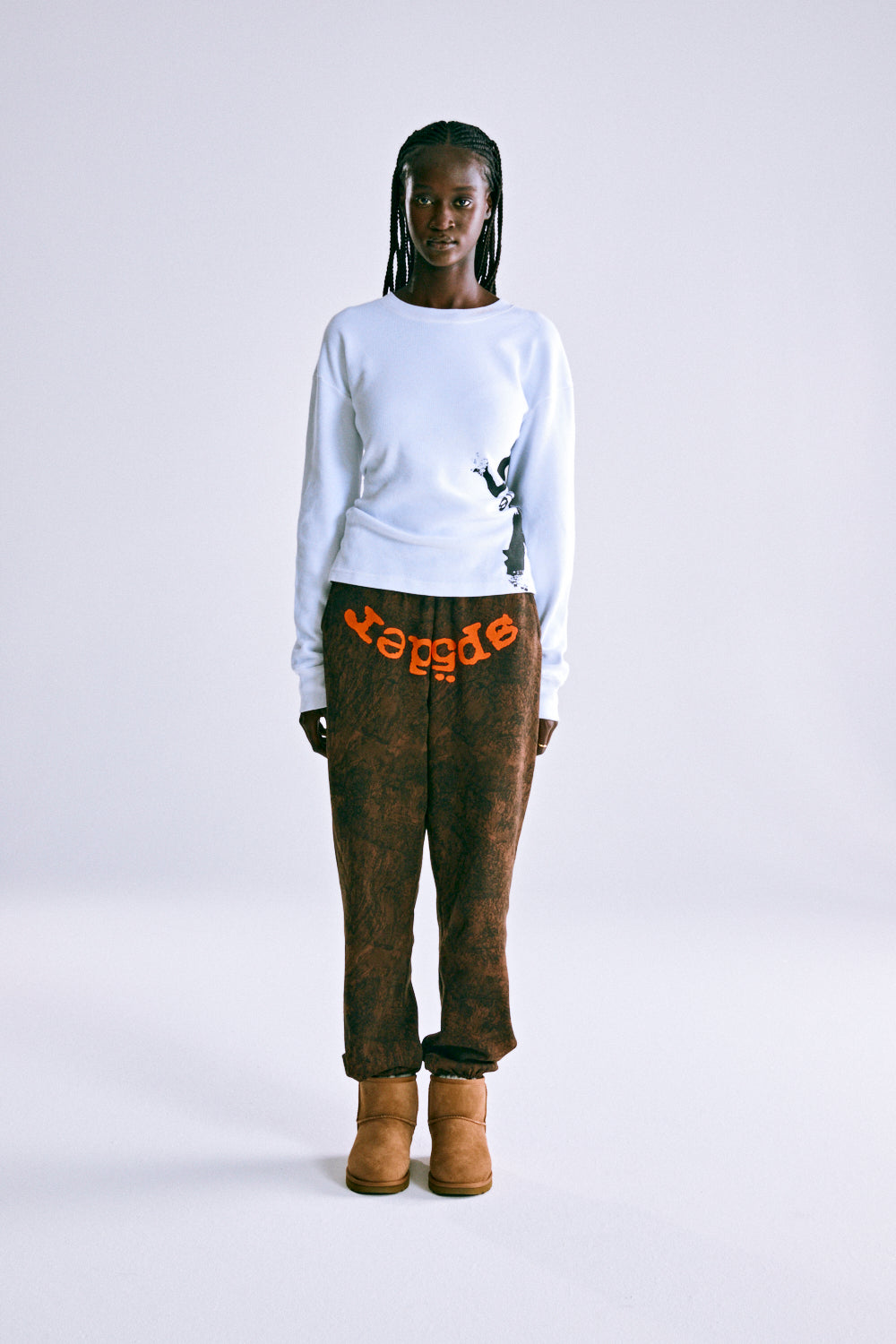 Model wearing brown tree camo sweatpants with orange SP5DER text and a white long-sleeve shirt.