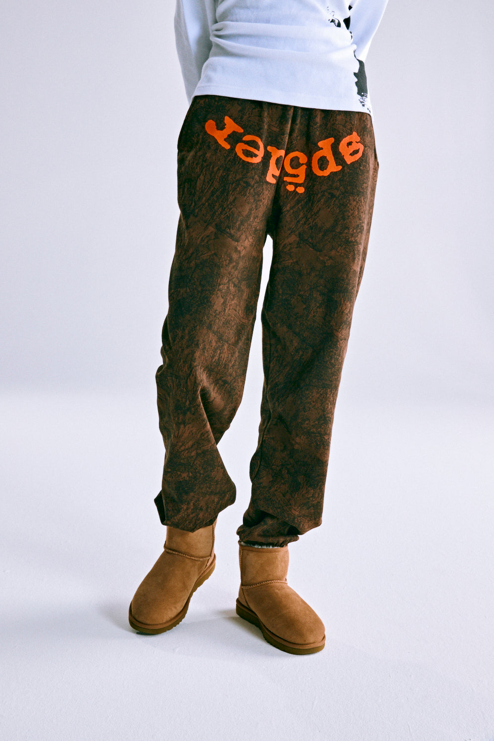 Brown tree camo sweatpants with orange "SP5DER" text, elastic cuffs, and drawstring waist.