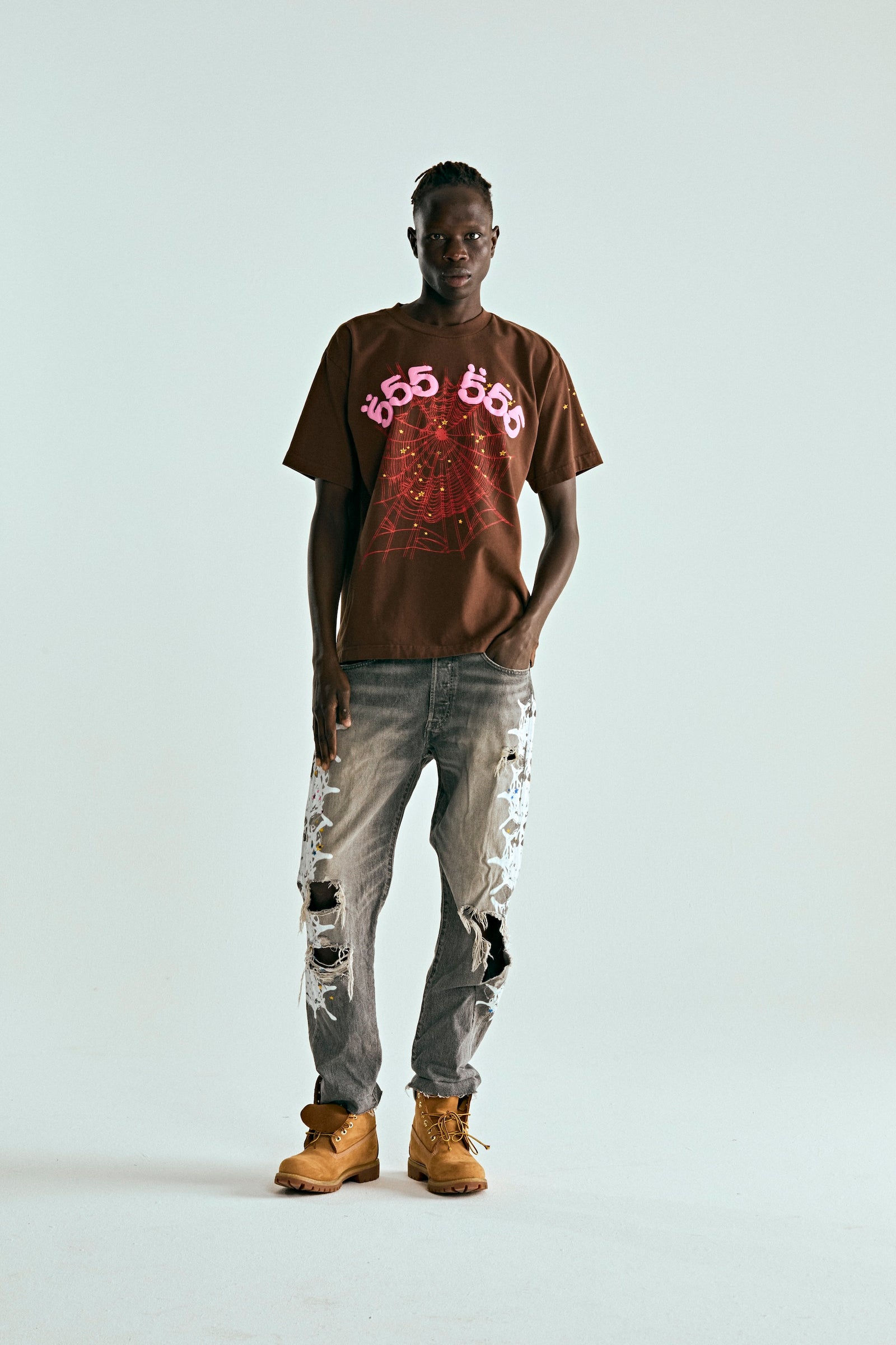 Brown ANGEL NUMBER TEE with pink "555" and red web print, worn by a man with distressed jeans.