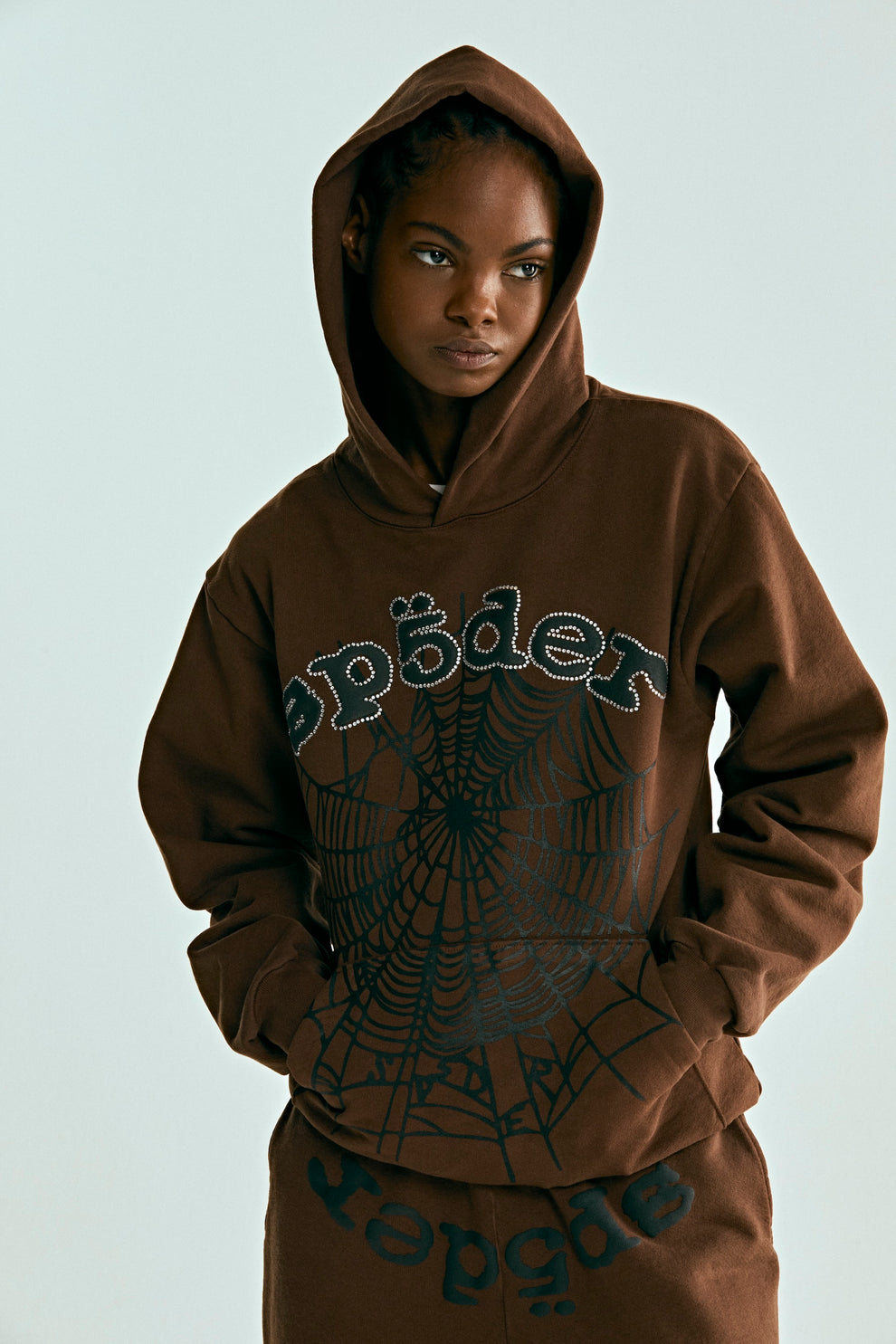 Brown OG WEB HOODIE with rhinestone "SP5DER" text and black spiderweb print, worn by a model.