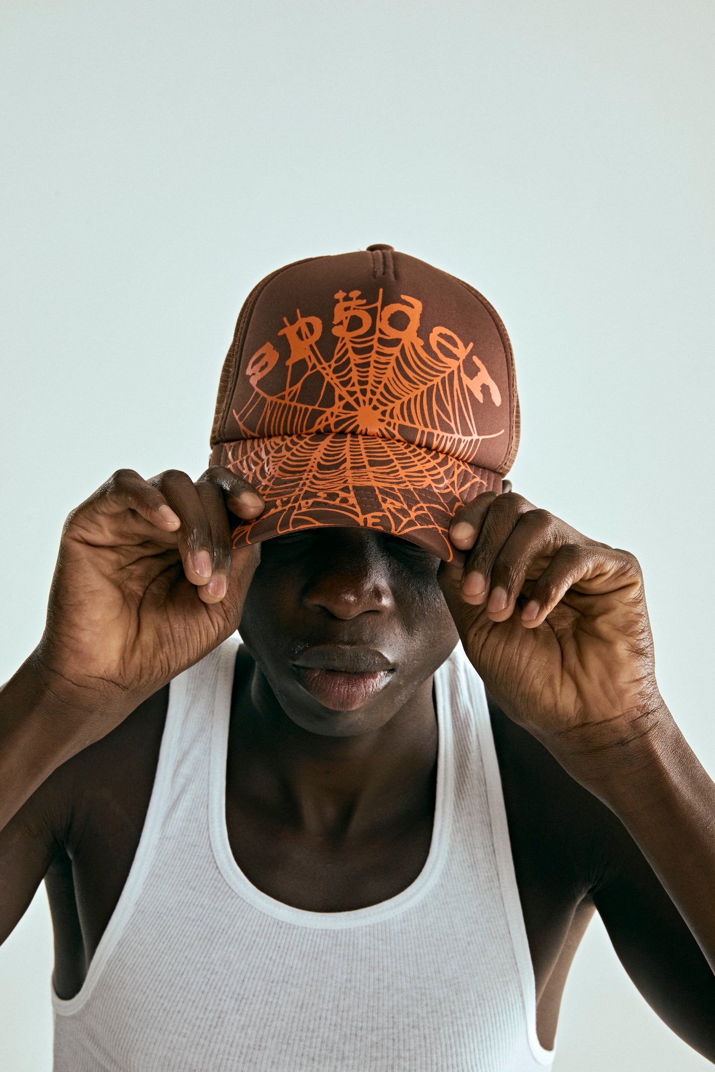 OG WEB V2 TRUCKER hat: brown curved brim cap with orange spider web print, worn by a person in a white tank top.