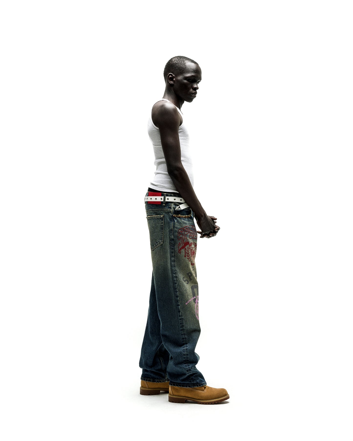 Man in baggy blue jeans with red and pink graffiti-style prints, white tank top, and tan boots.