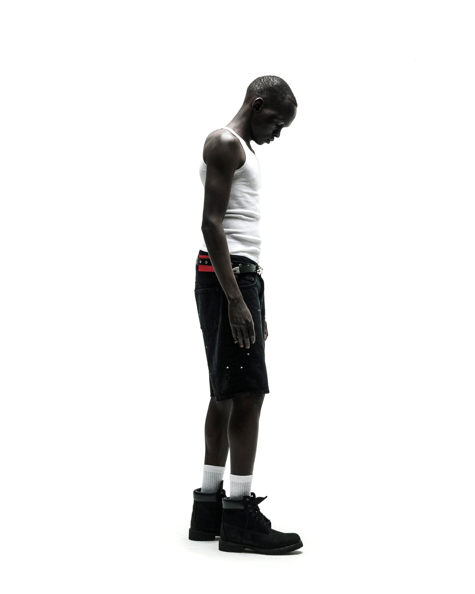 Man in black denim jean shorts, white tank top, red belt, and black boots.
