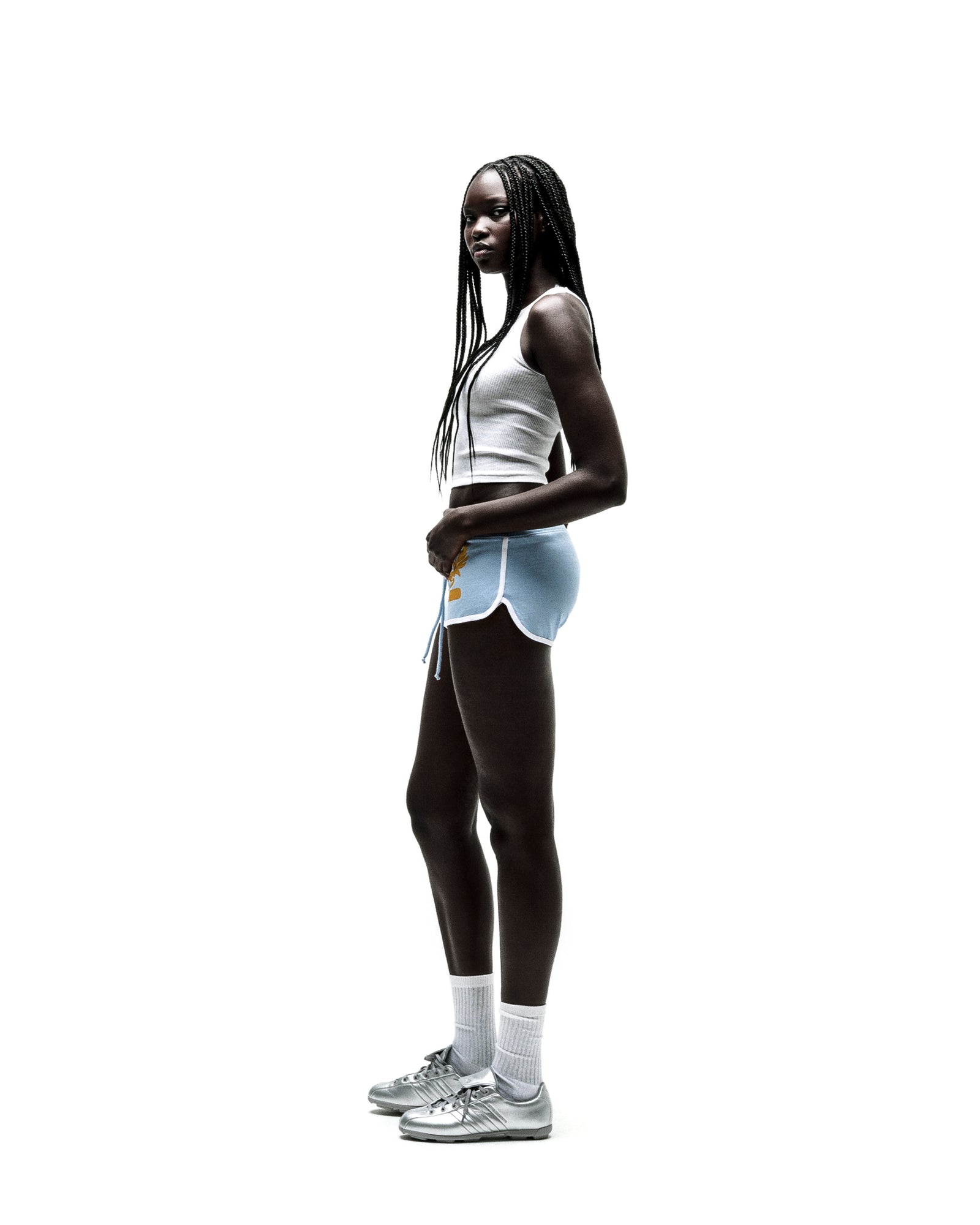 Young woman in light blue SP5 WIDE BOXER shorts with white trim, a white crop top, and silver sneakers.