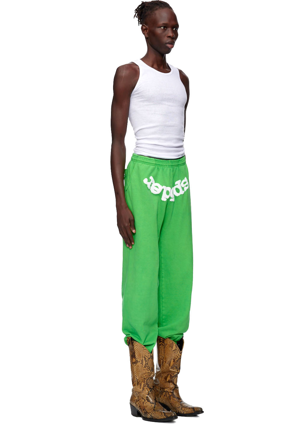 Model wearing green CLASSIC SWEATPANTS with white puff print "SP5DER" and web print, paired with a white tank top and snakeskin boots.