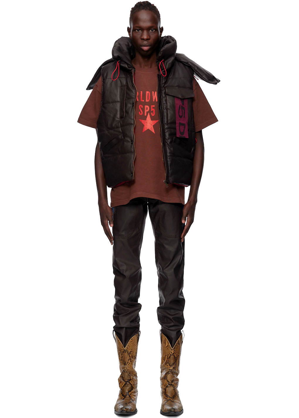 Man wearing a brown coated canvas hooded puffer vest with red accents over a brown graphic tee and dark pants.
