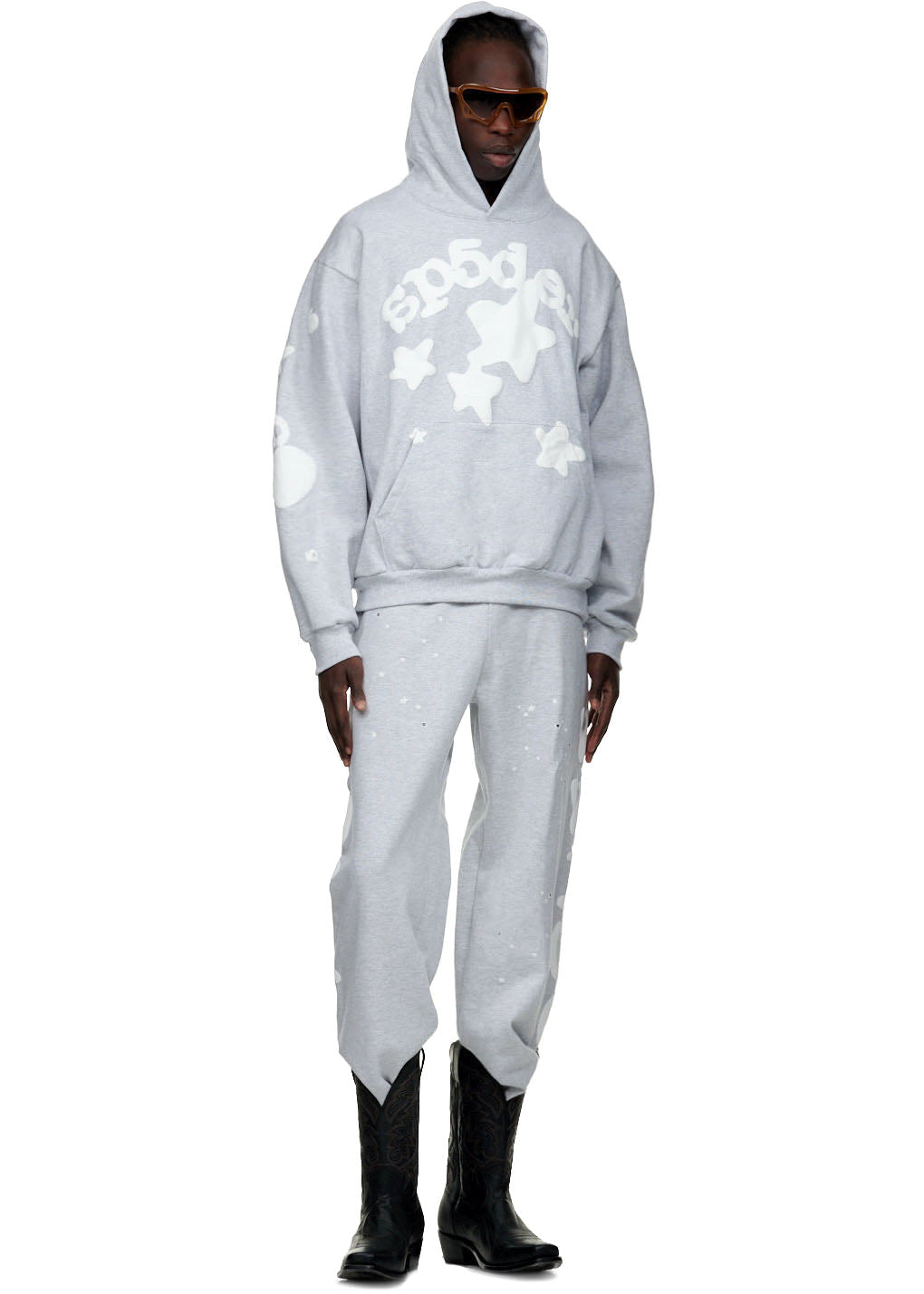 Male model in grey Beluga sweatpant and matching hoodie with white star puff prints, wearing black cowboy boots.